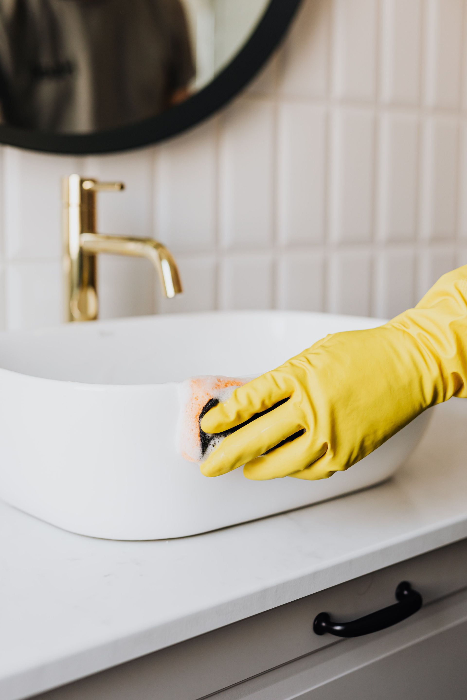 Crop faceless person in yellow rubber protective glove wiping surface of white sink in bathroom with sponge and cleaning agent